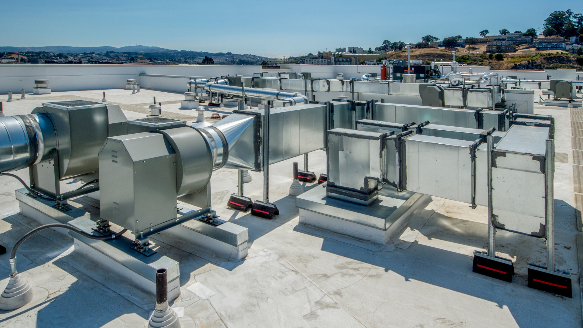 Rooftop HVAC system with metal ductwork and fans on a flat white roof against a clear blue sky.