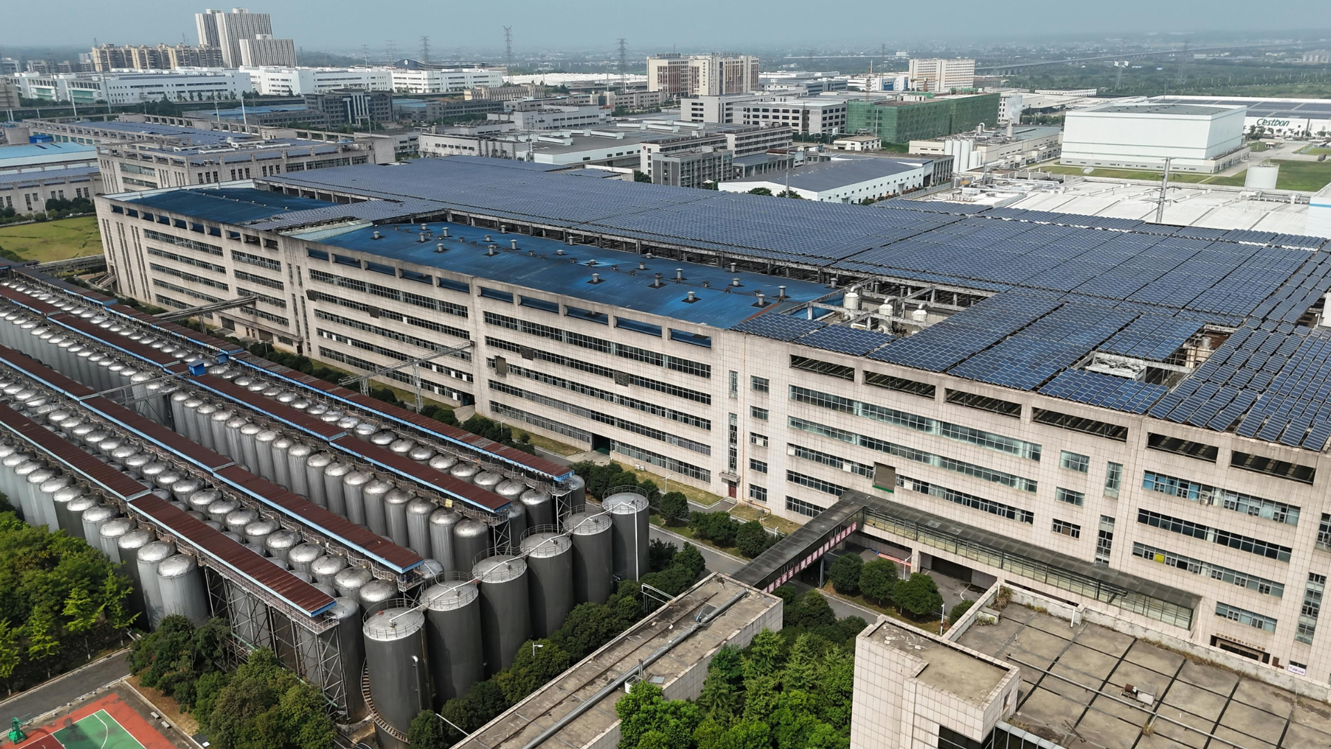 An aerial view of a large industrial factory complex with extensive rooftop solar panels and silver storage silos.