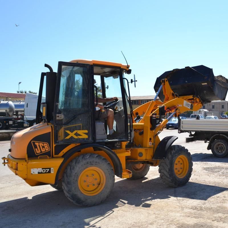 Pala caricatrice JCB gialla con benna sollevata, parcheggiata sul cemento. Cabina aperta. Giornata di sole.