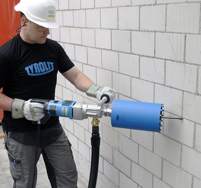 Uomo che pratica un foro in un muro di mattoni bianchi con un trapano a carotaggio, indossando dispositivi di sicurezza.