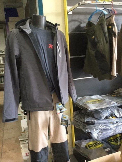 Manichino in abbigliamento outdoor: giacca grigia, camicia blu, pantaloni beige e neri. Espositore da negozio con pile di vestiti.