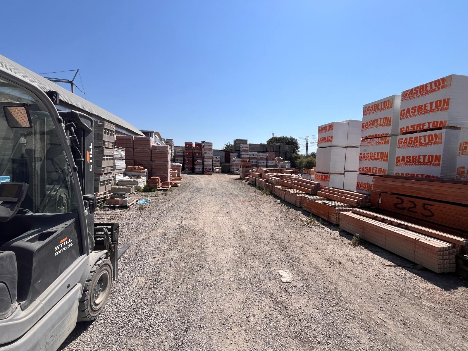 Carrello elevatore in un deposito di ghiaia pieno di pile di mattoni e materiali da costruzione sotto un cielo azzurro e limpido.