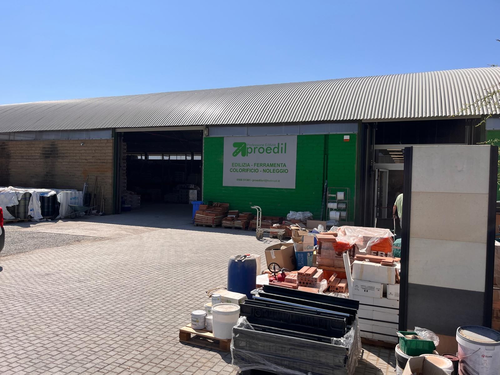 Esterno di un negozio di forniture edili, prodotti esposti all'ingresso. Il negozio ha un'insegna verde e il pavimento è asfaltato.