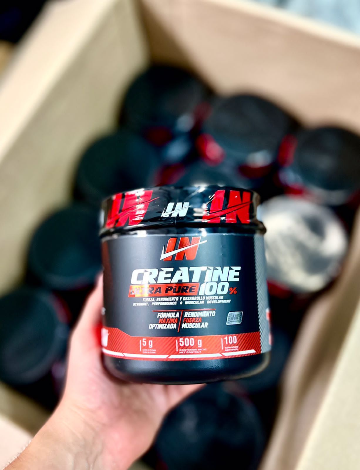 Una mano sostiene un bote de creatina en polvo de color negro y rojo frente a varios botes similares.