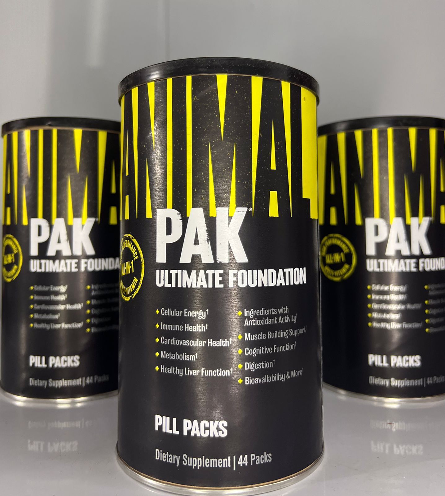 Tres envases de base de maquillaje AKMA PAK de color negro y amarillo sobre una superficie reflectante.