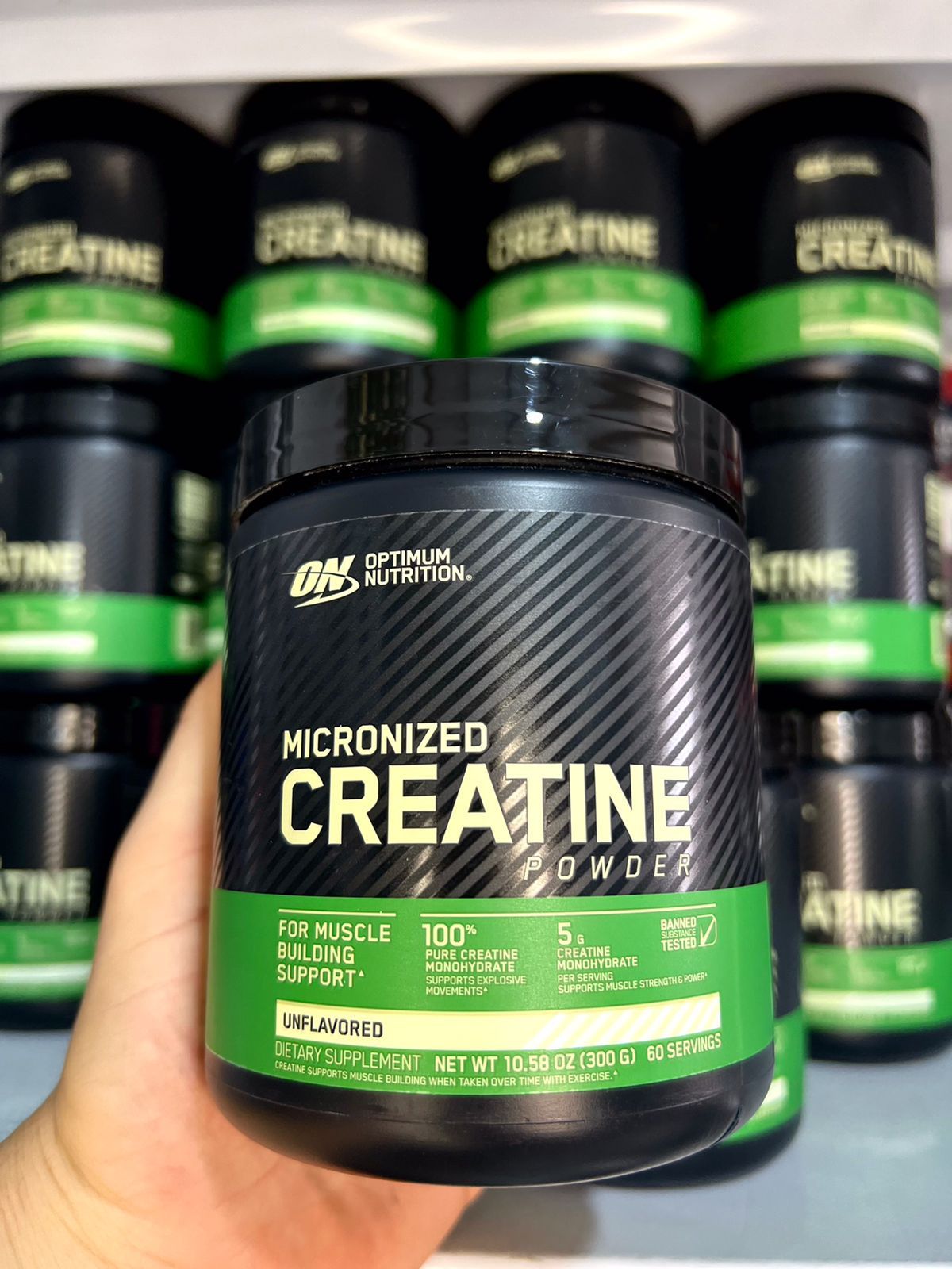 Una mano sostiene un recipiente negro verdoso de creatina micronizada frente a una pila de envases de creatina.