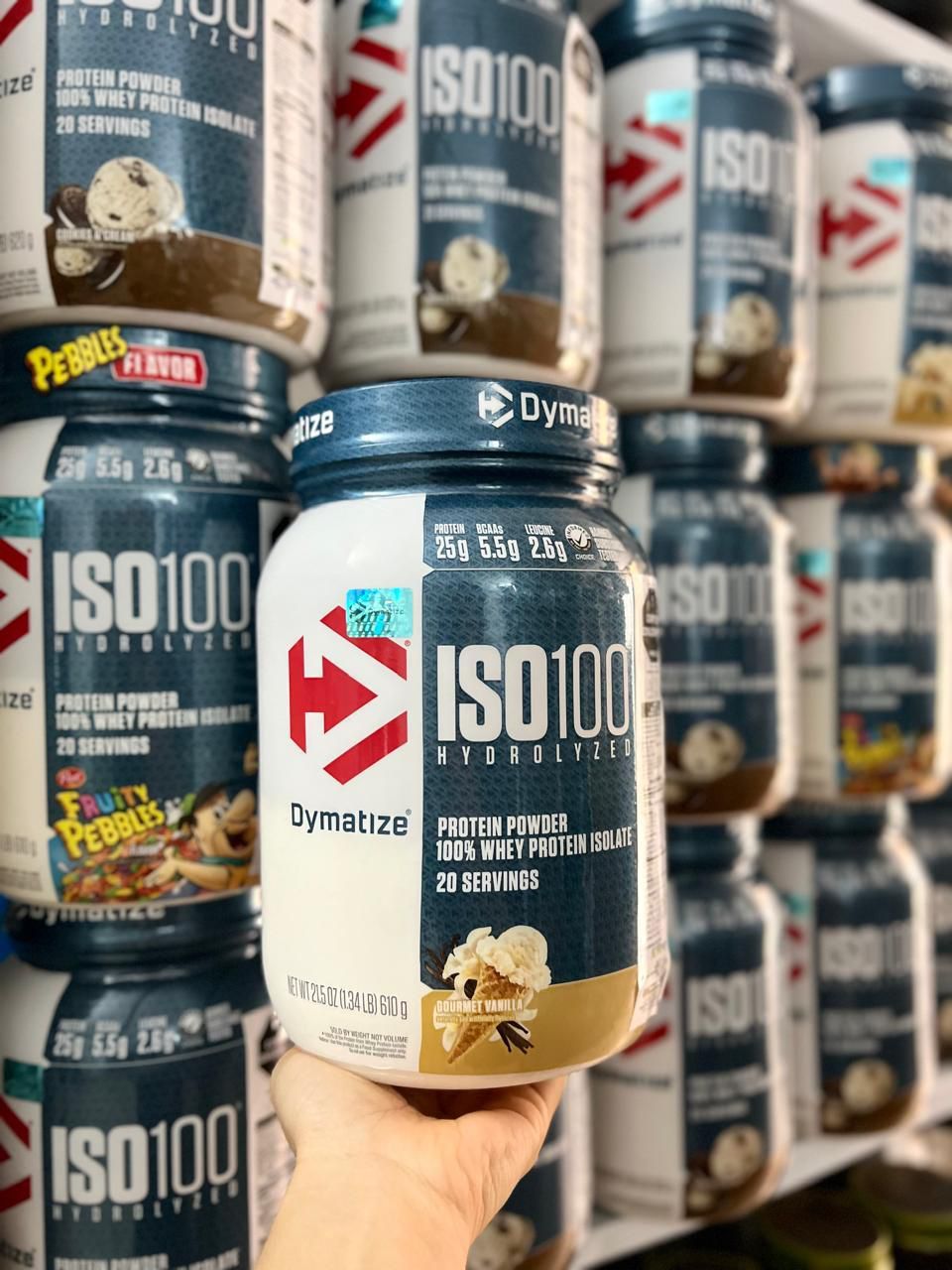 Una mano sostiene un bote de proteína en polvo “ISO100”, con botes apilados al fondo.