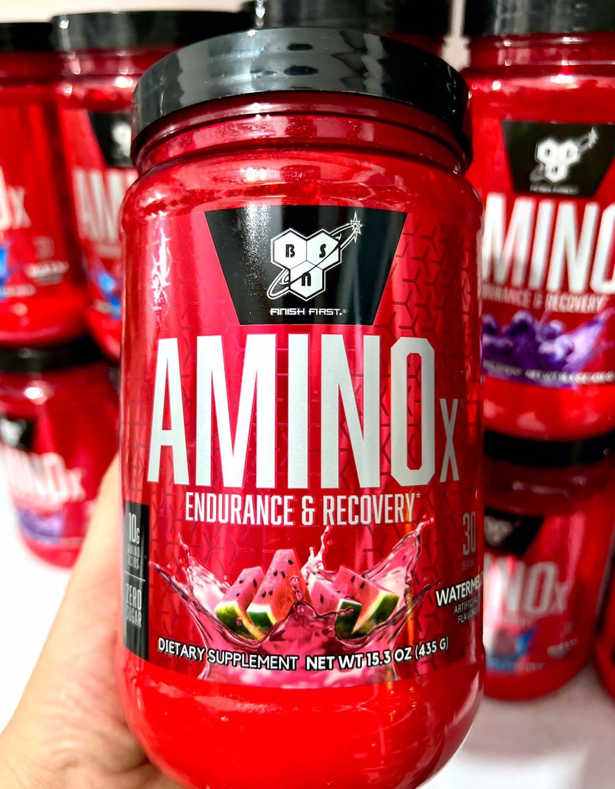 Frasco de suplemento Red Amino X con etiqueta blanca en un estante de tienda, rodeado de más frascos rojos.