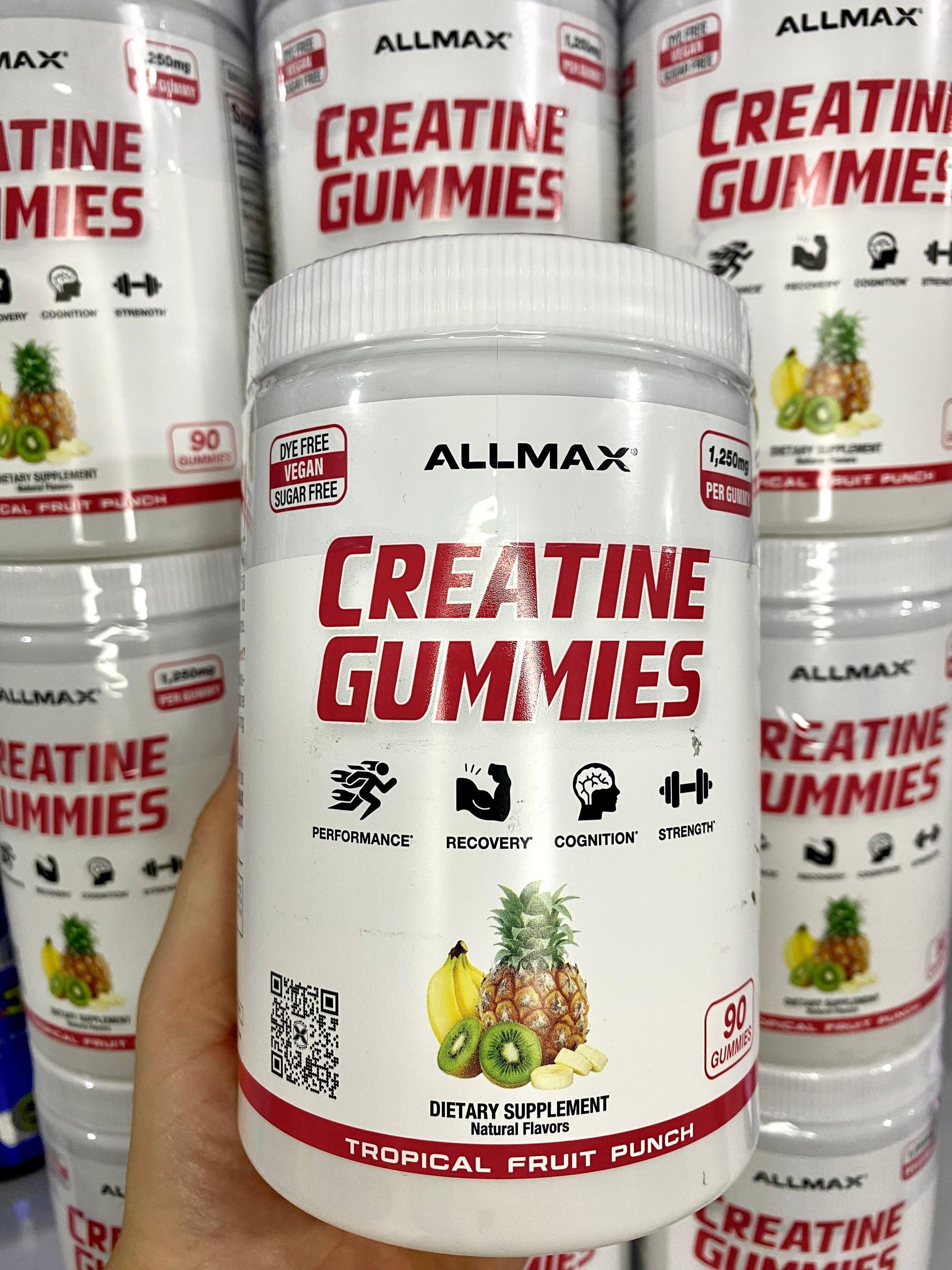 Frascos de gomitas de creatina Allmax en envases blancos y rojos, un frasco sostenido frente a varios otros.