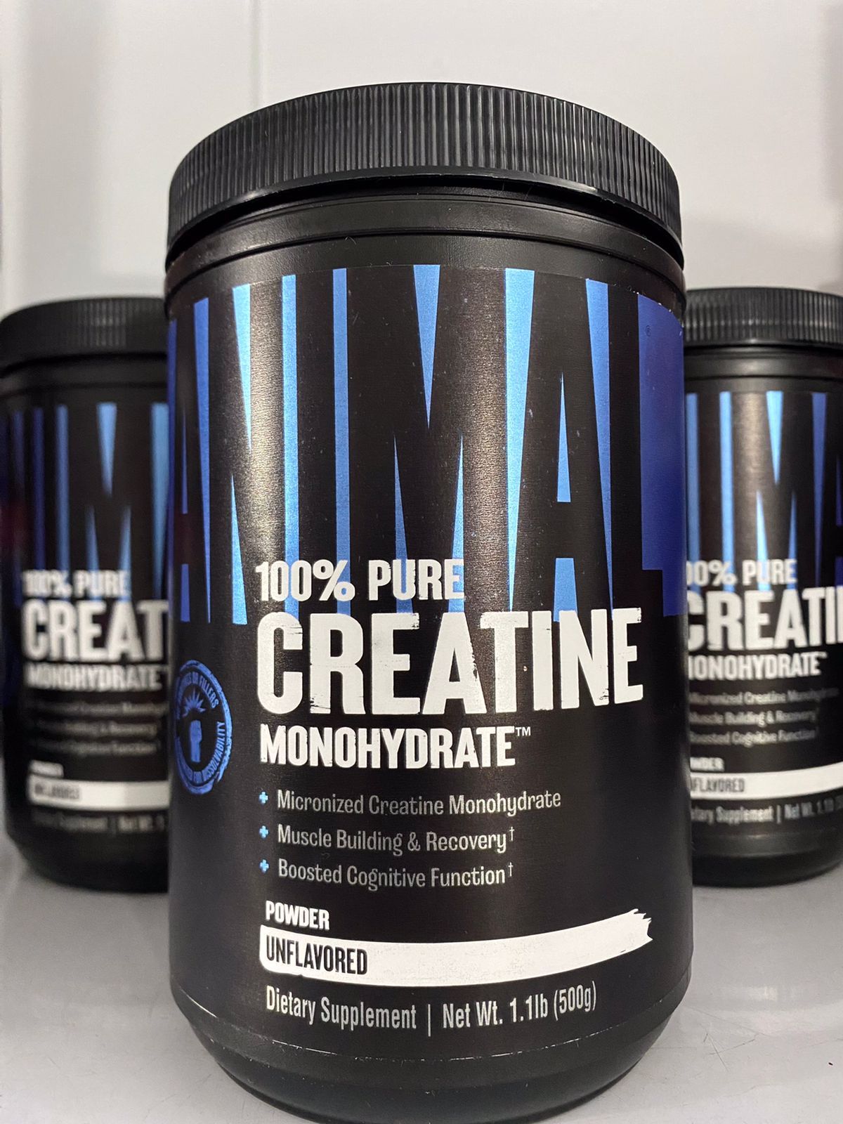 Frasco negro de suplemento de monohidrato de creatina con detalles azules, flanqueado por dos envases similares.