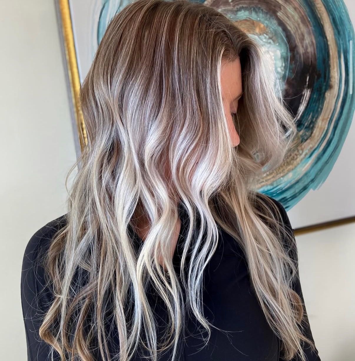 Woman with blonde highlights, wearing black top, posing near abstract art.