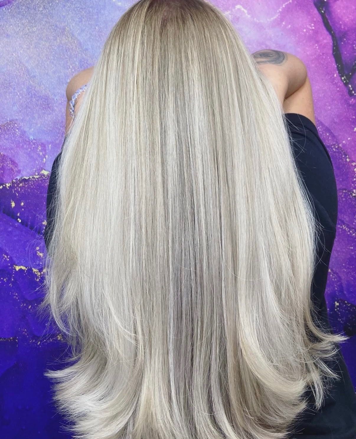 Long, light blonde hair cascading down the back; a person in dark clothing sits against a purple backdrop.