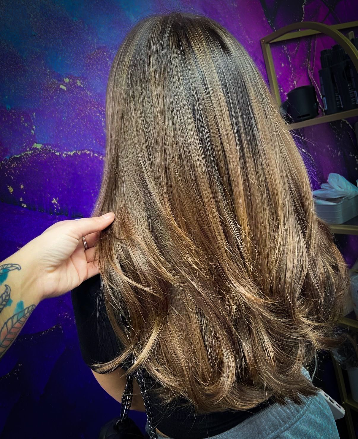 Brown hair with highlights, shoulder-length, being touched by a hand with tattoos in front of a purple backdrop.