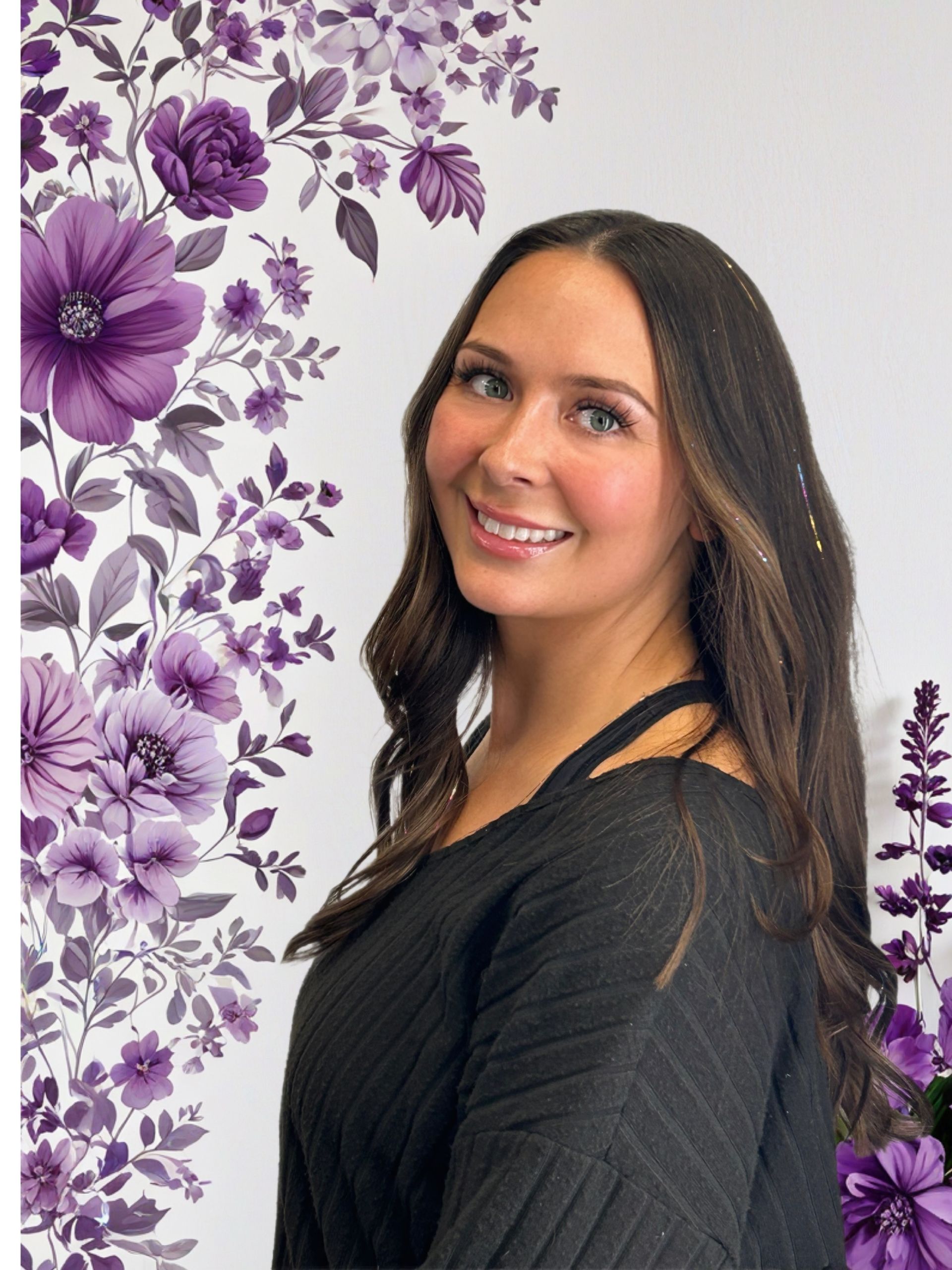 Maddy with long brown hair smiles, wearing a black top, in front of a purple floral background.