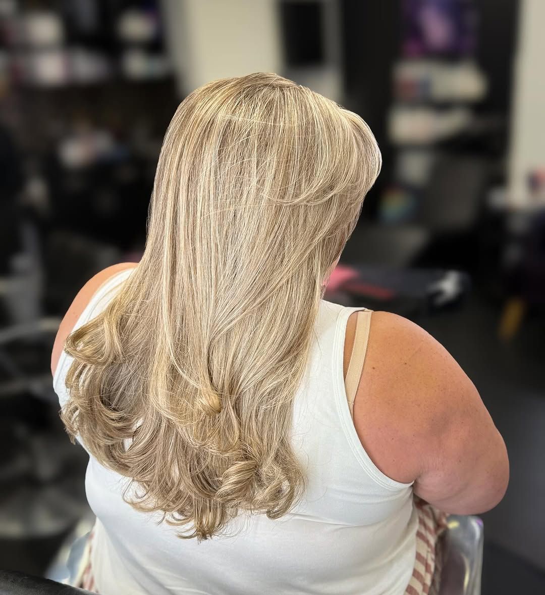 Woman with long, layered blonde hair, wearing a white tank top in a salon.