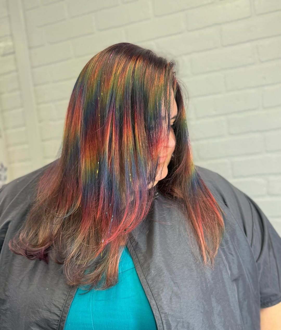 Person with long, rainbow-colored hair, wearing a gray salon cape, in front of a white brick wall.