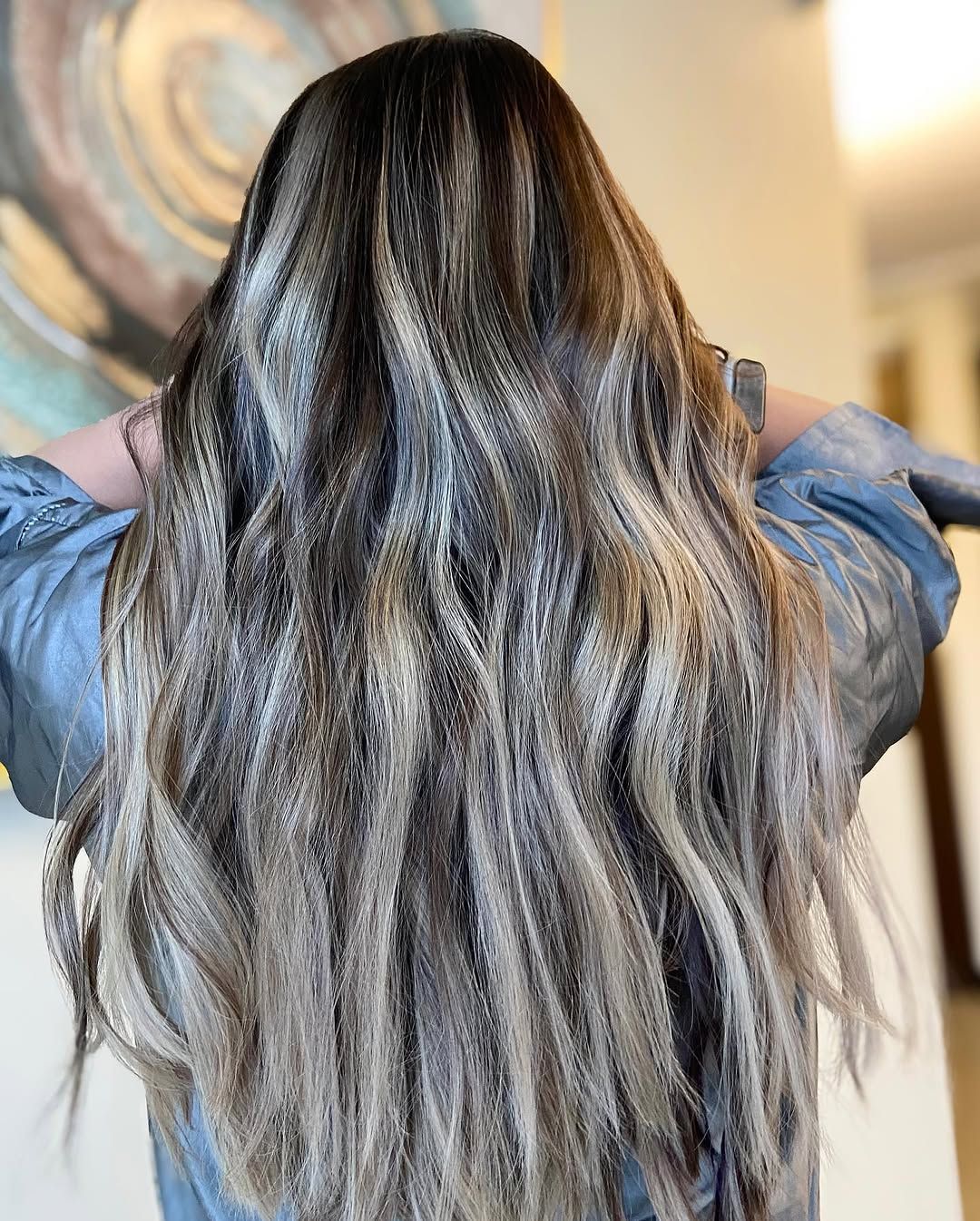 Woman with long, wavy hair, dark roots and light brown and blonde highlights, seen from behind.