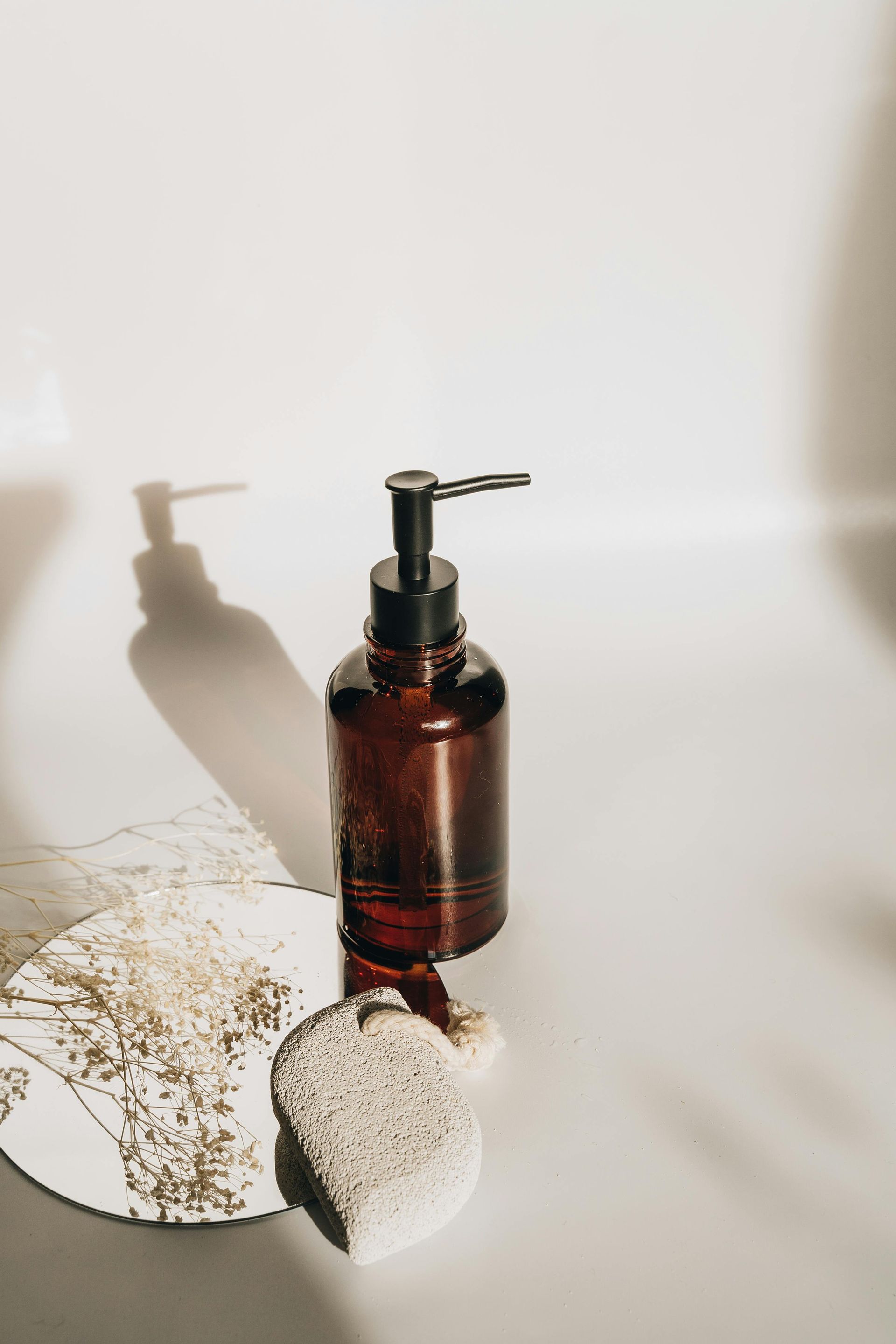 Amber bottle with black pump dispenser on a white surface, casting a long shadow, next to a loofah.
