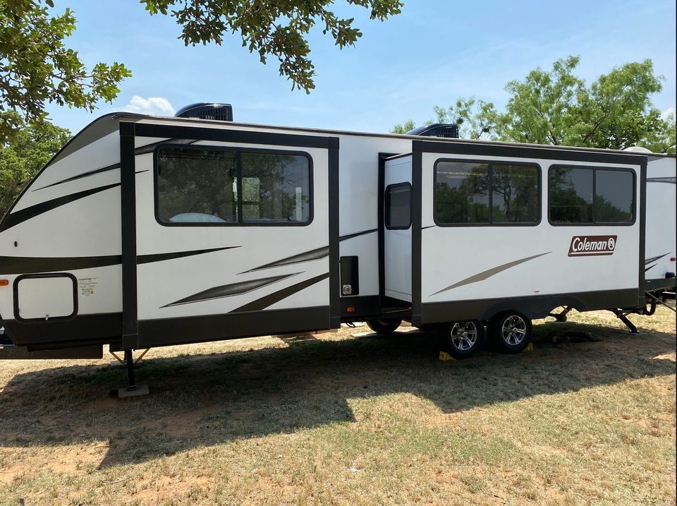 White and black Coleman travel trailer parked on grass under a clear blue sky.