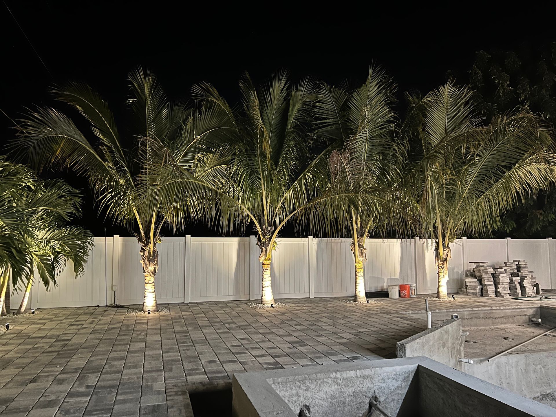 Palm trees illuminated at night, in front of a white wall, with a paver patio and unfinished concrete structure.