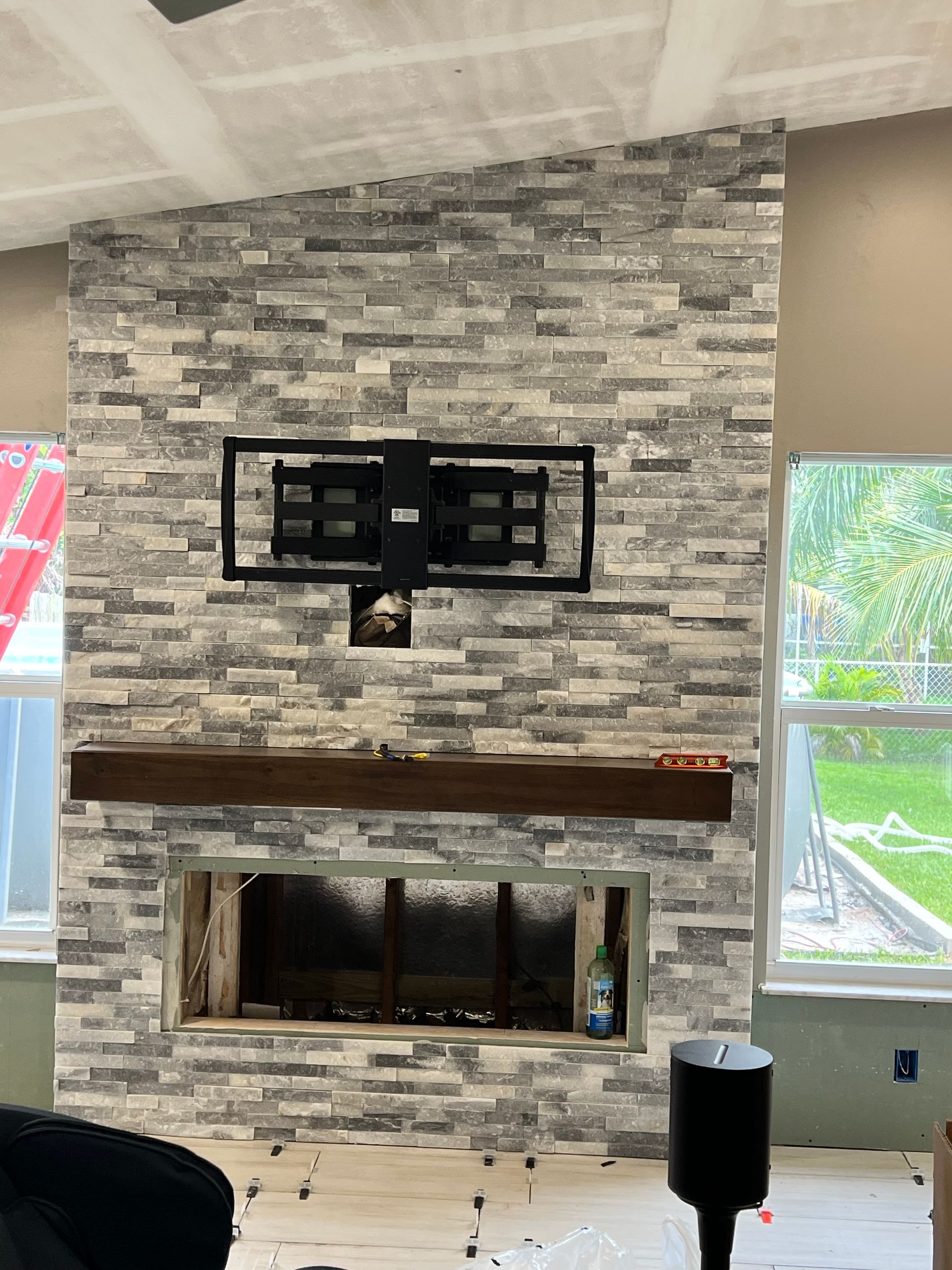 Fireplace with gray and white stone, TV mount, wooden mantel, and windows in a room under construction.