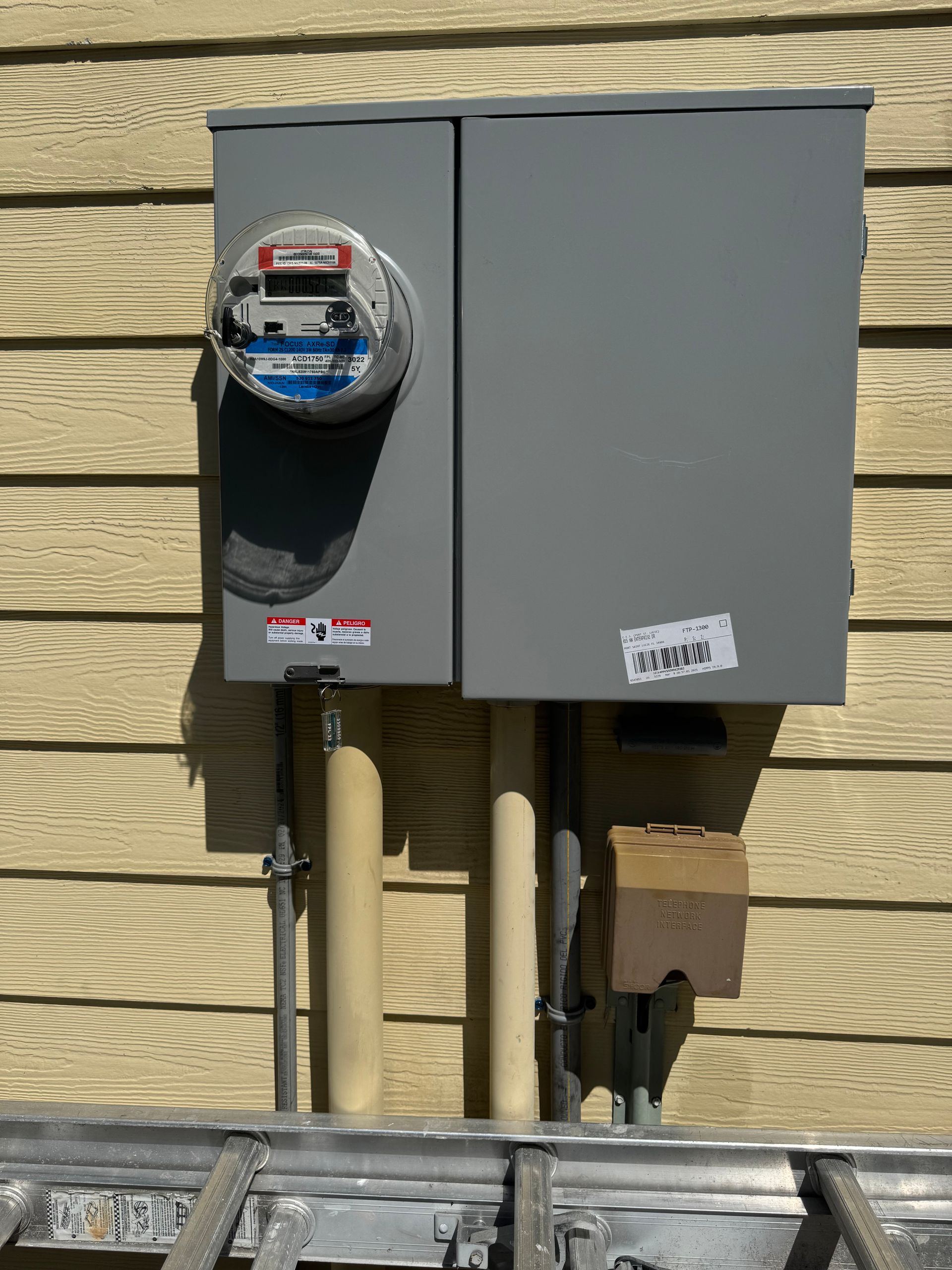Electric meter and panel mounted on a yellow-sided building. Conduit and ladder in view.