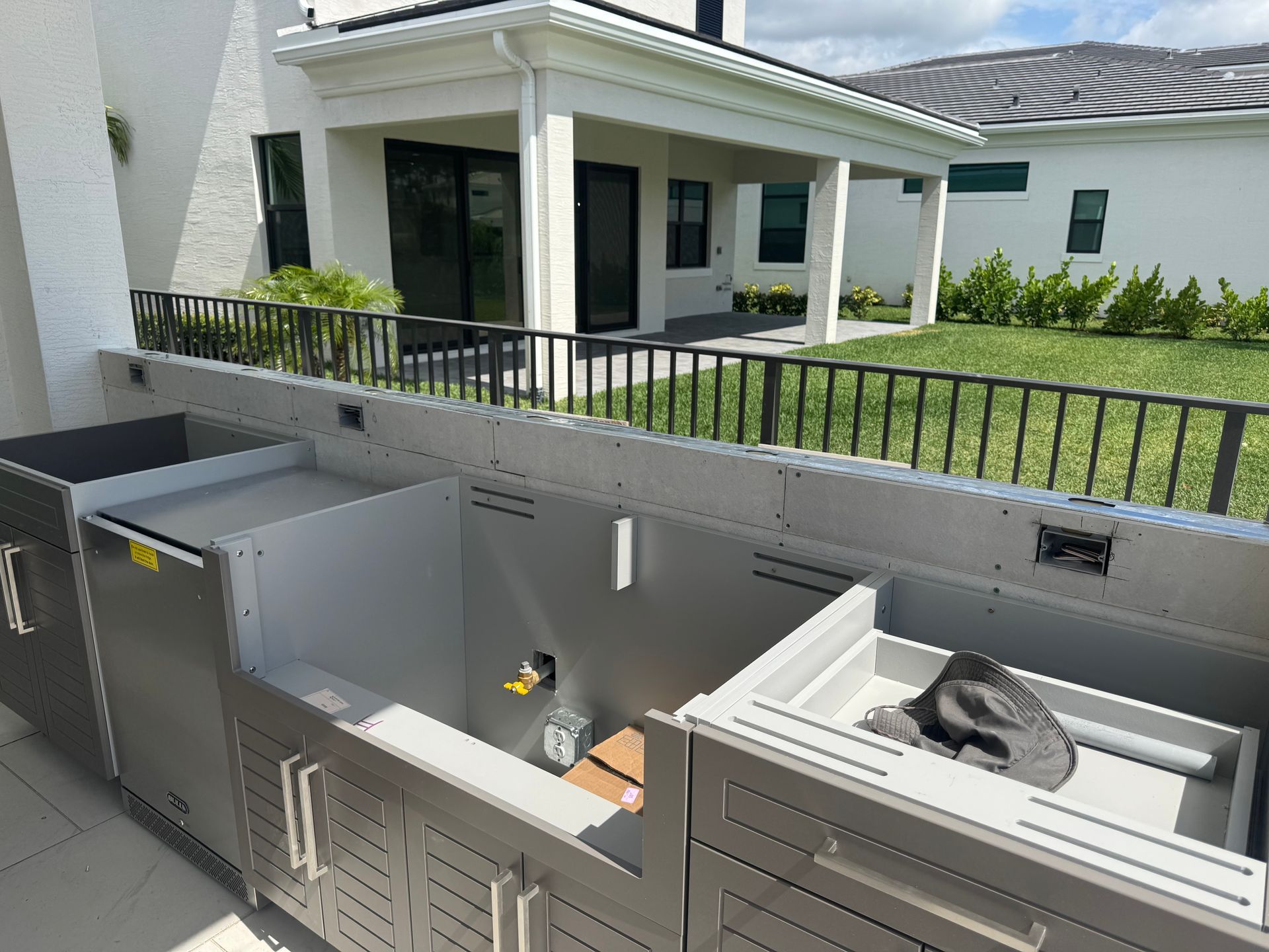 Outdoor kitchen cabinets, stainless steel, under a patio roof, with a fence and house in the background.