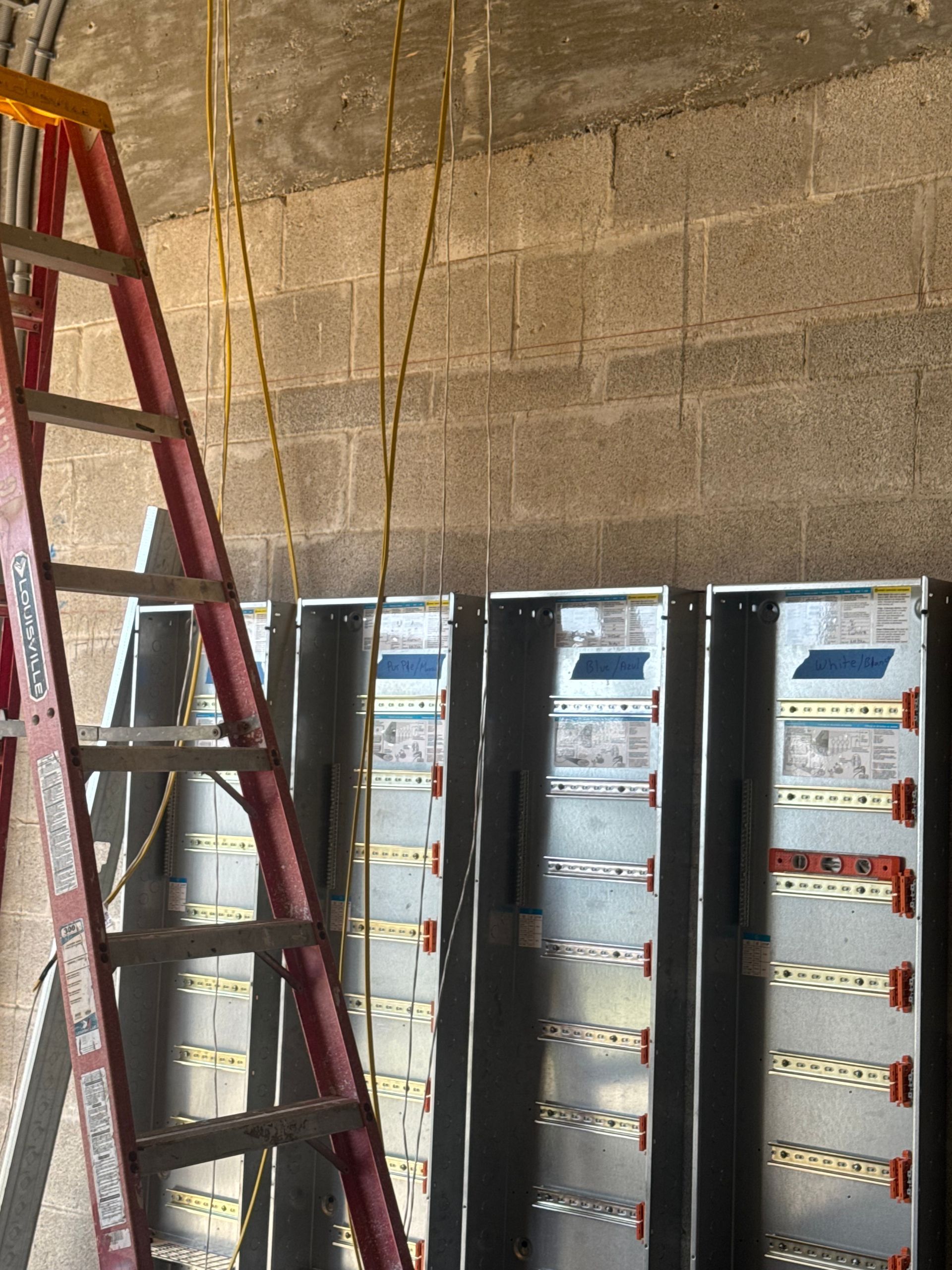 Red ladder next to three electrical panel boxes mounted on a gray cinder block wall.