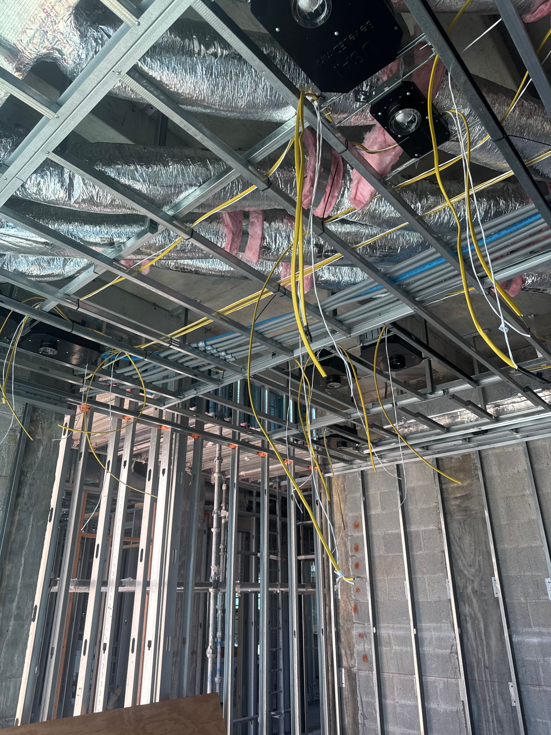 Construction worker in a yellow hard hat examines wiring in a wooden framed building.