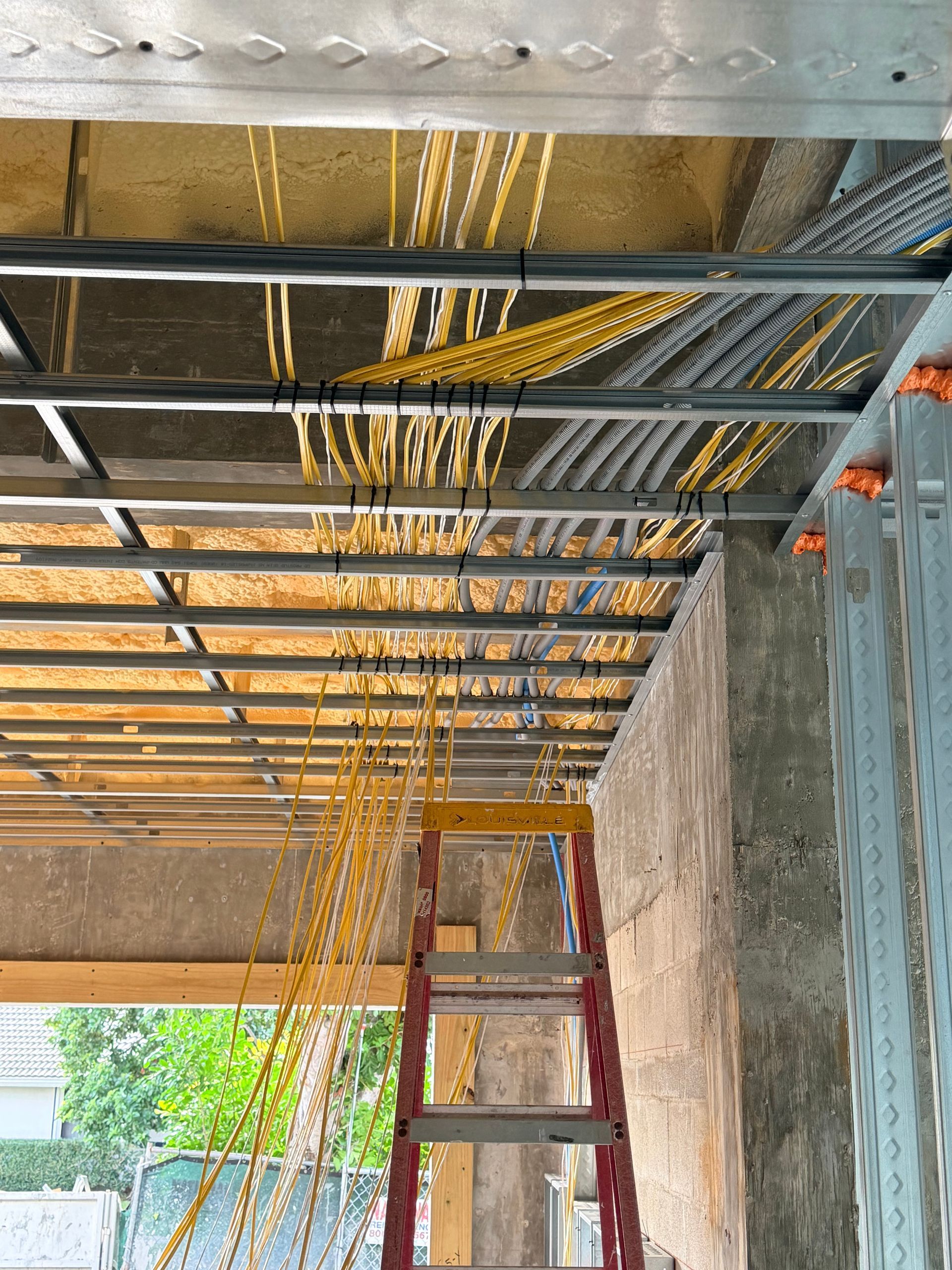 Yellow electrical wires hanging from a metal framework ceiling, construction site interior. A red ladder stands below.