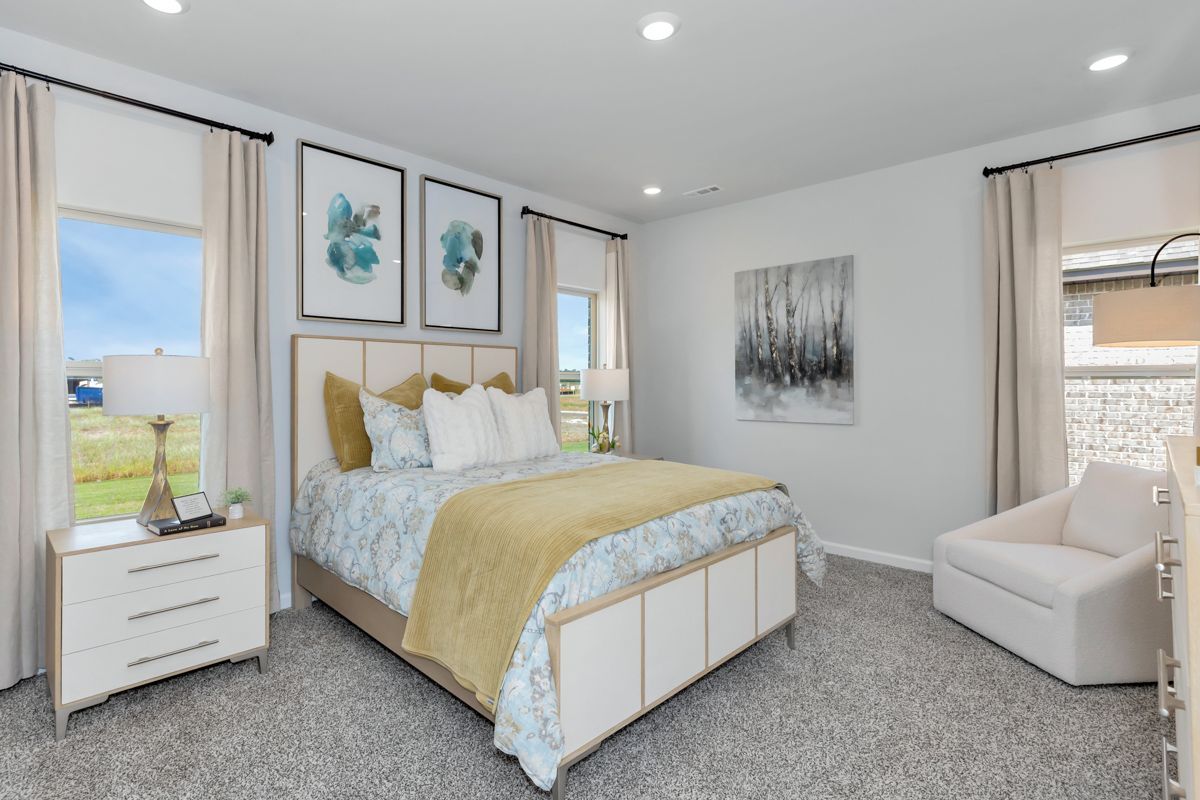 A bedroom with a bed, chair, nightstand, two windows, and artwork. The walls are white.