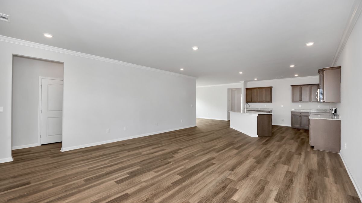 Empty open-concept living space with wood floors, white walls, and a kitchen in the background.