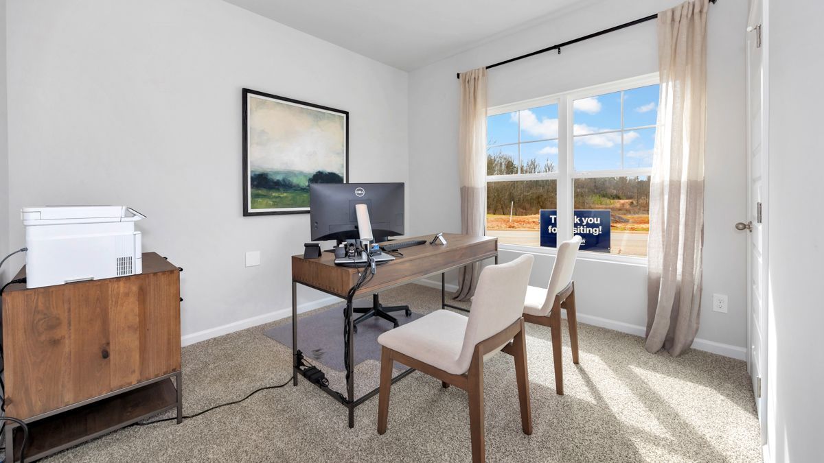Home office with desk, chairs, and window view. Light gray walls, neutral carpet, and a wooden cabinet.