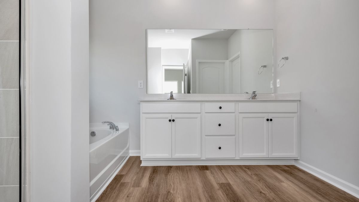 White bathroom with double vanity, large mirror, and wood-look flooring. Bathtub visible on the left.