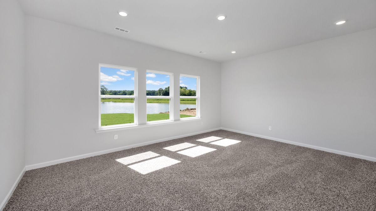 Empty room with gray carpet, white walls, and a window overlooking a lake. Sunlight shines in.
