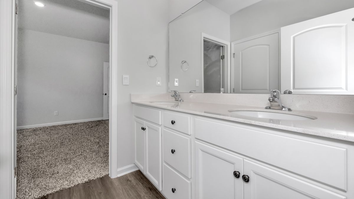 Bathroom with white cabinets, double sinks, and a doorway to a bedroom with carpet.