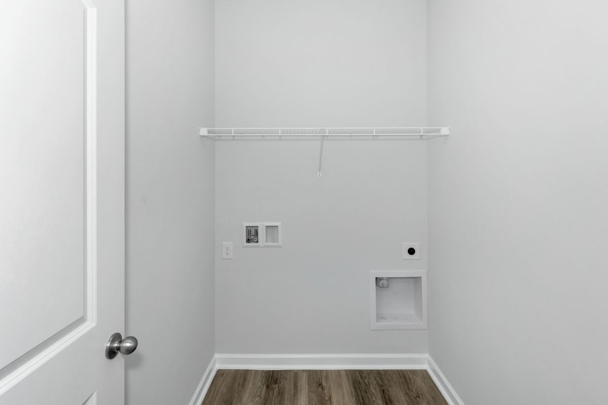 Empty laundry room with white walls, wood-look floor, and wire shelf.  Electrical and water hookups visible.