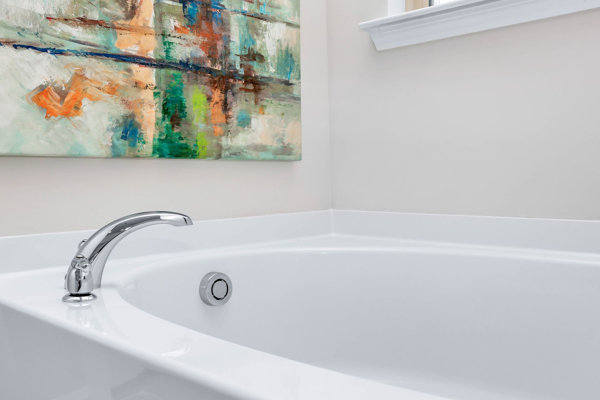 White bathtub with chrome faucet, in a bathroom with abstract art and a window.