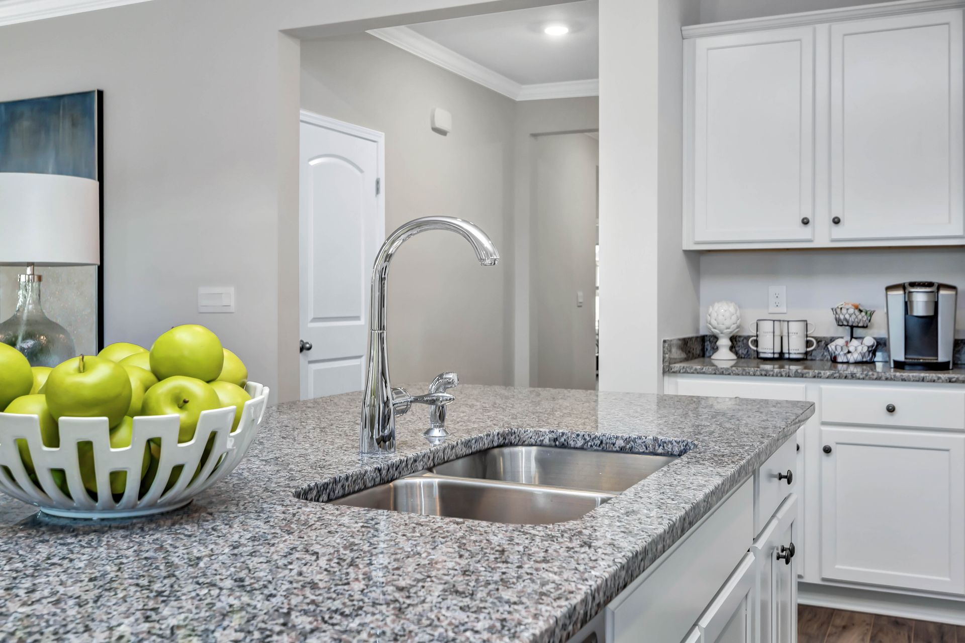 Kitchen with granite countertop, stainless steel sink, and white cabinets; bowl of green apples.