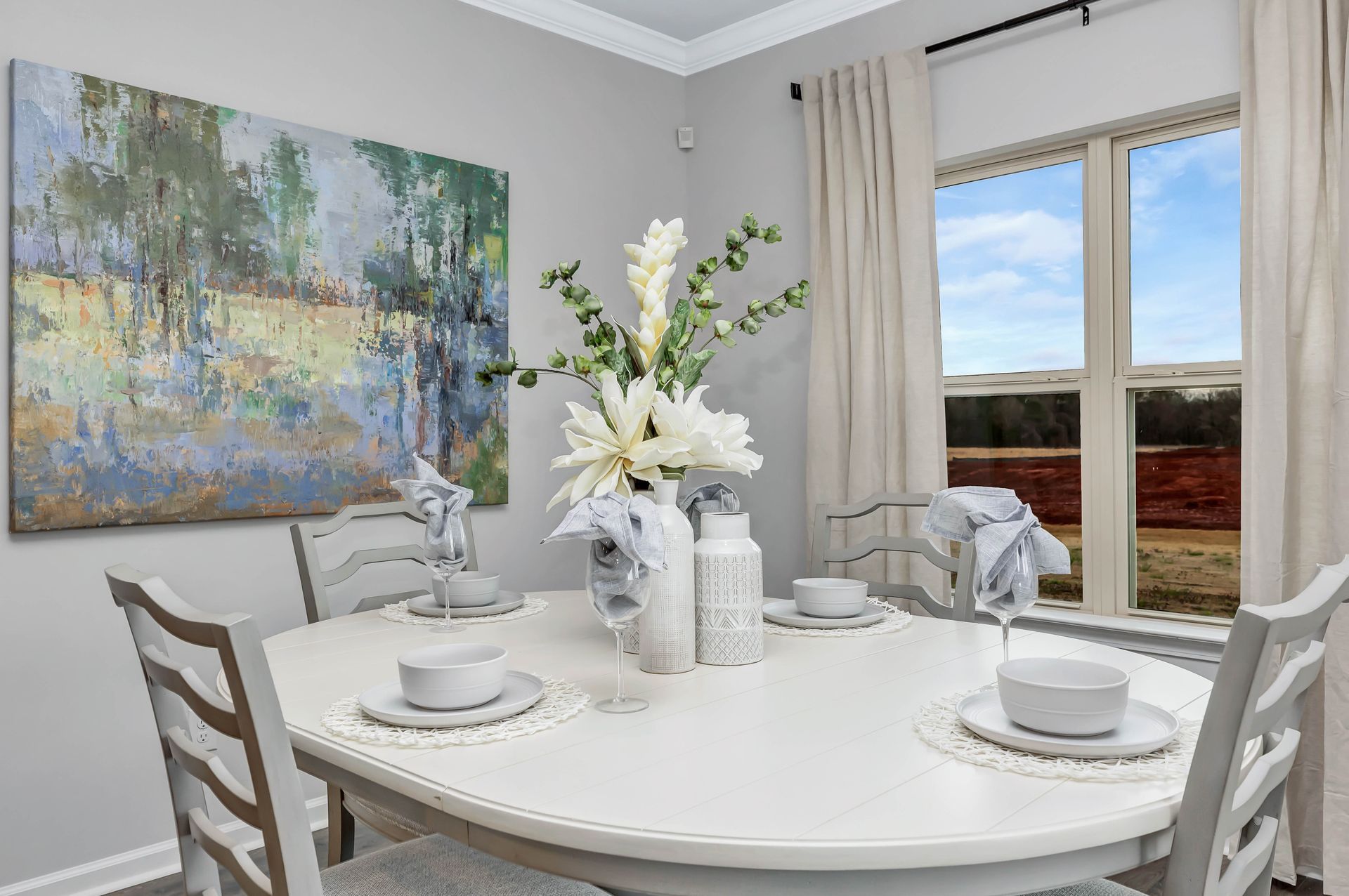 Dining room with white table set for four; artwork, window, and decorative flowers present.