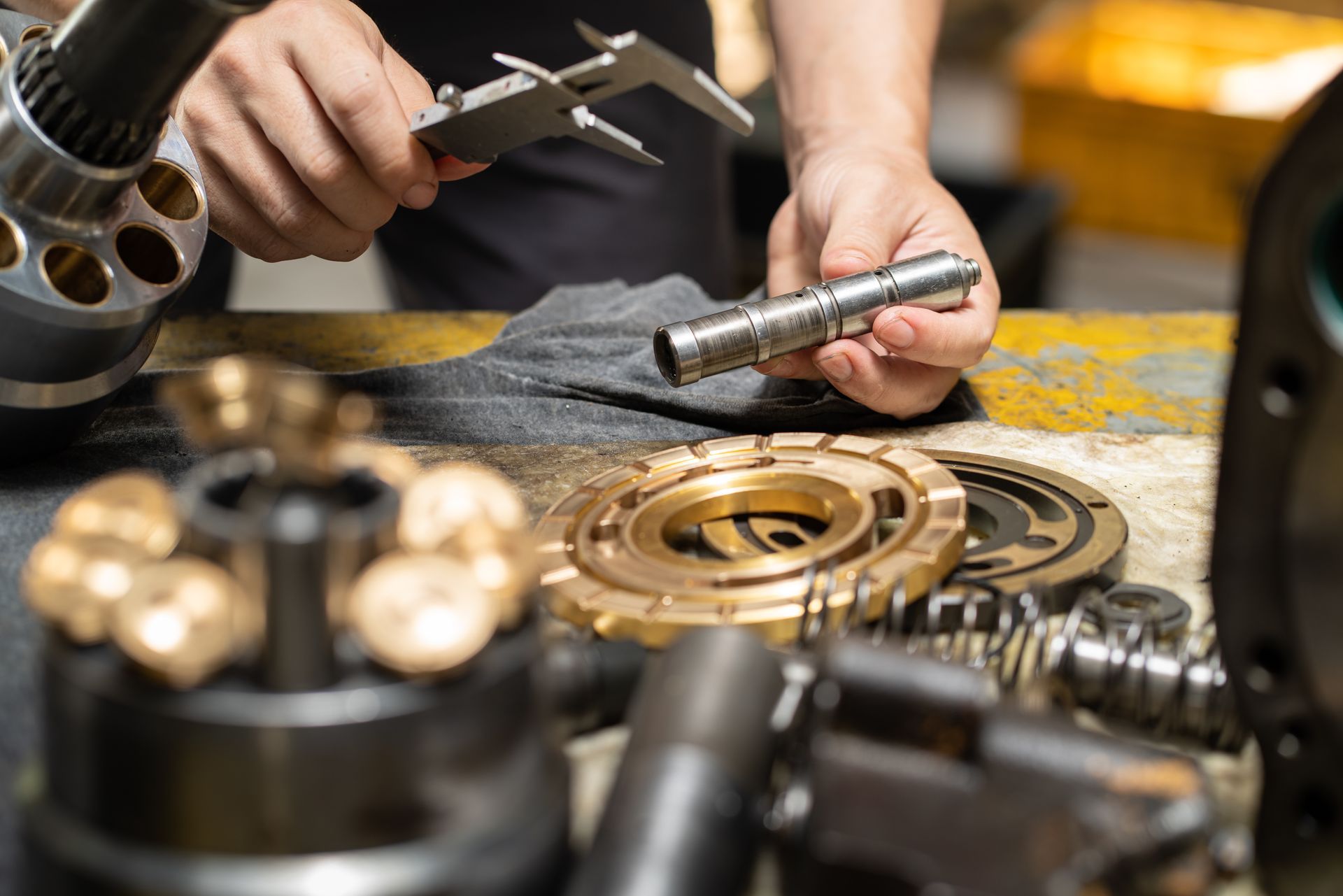 Hands measuring a metal shaft with calipers on a workbench with mechanical parts.