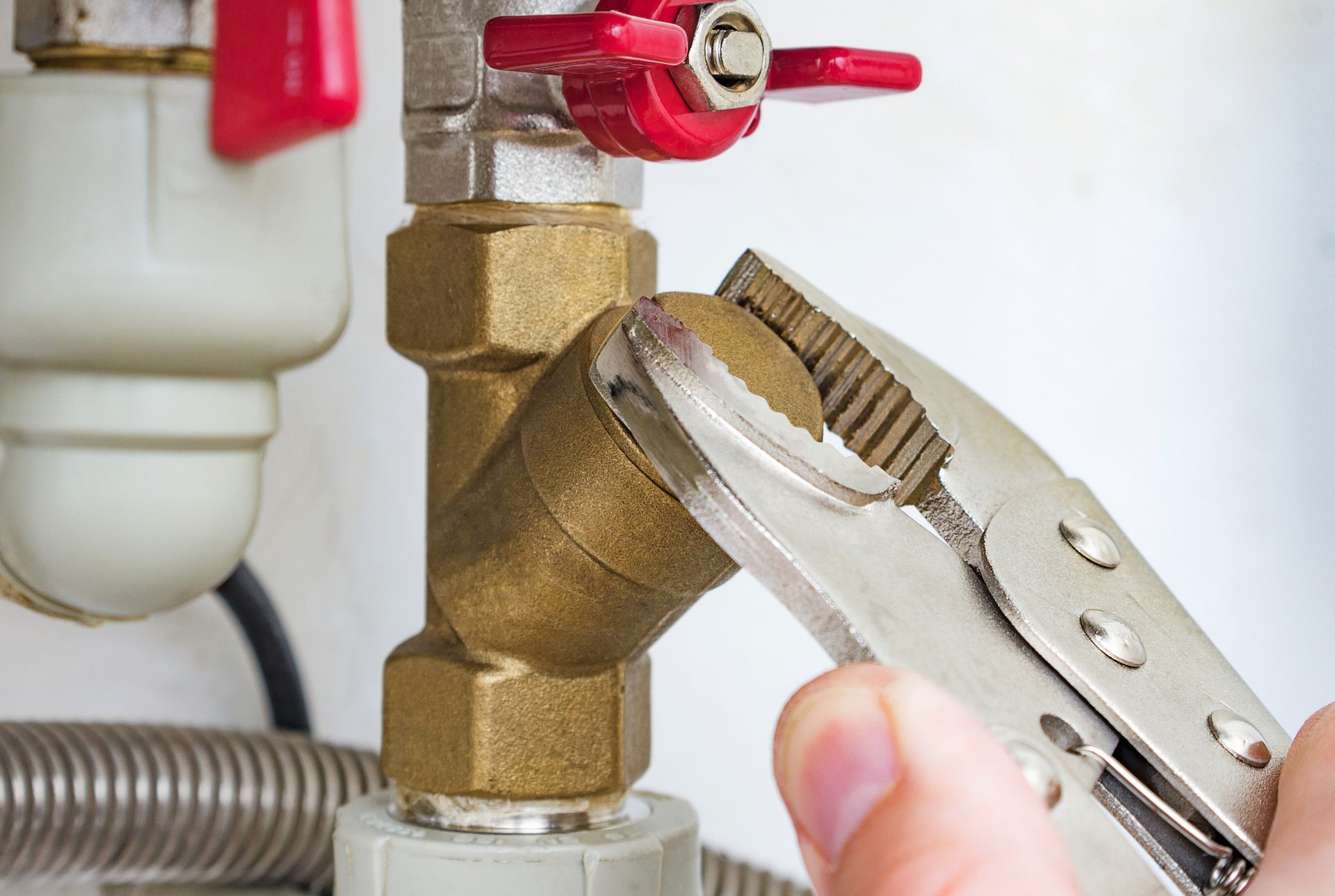 A hand using pliers to tighten a brass plumbing fitting with a red valve.