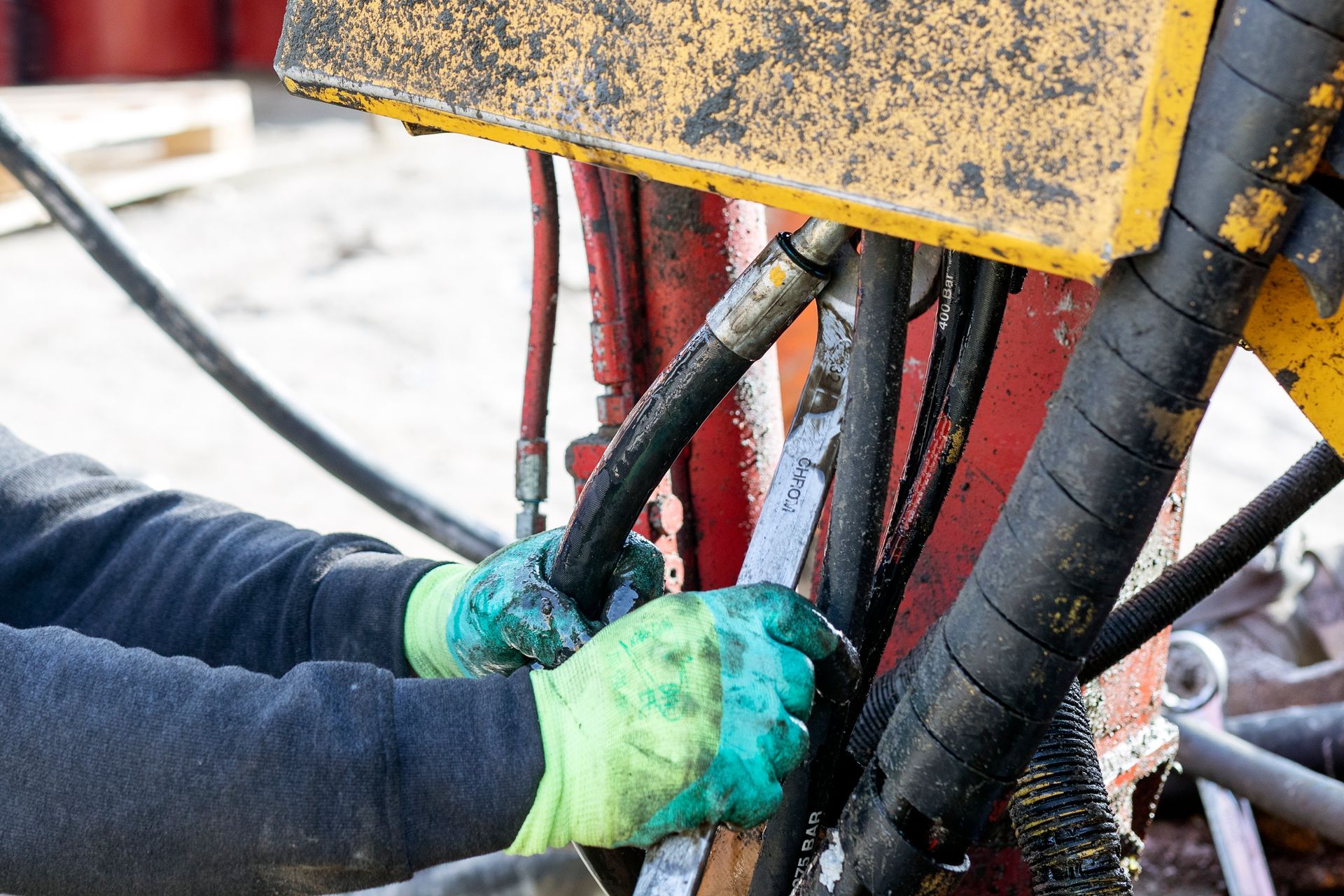 Person in green gloves uses a wrench on hydraulic hoses of a machine, likely for repair.