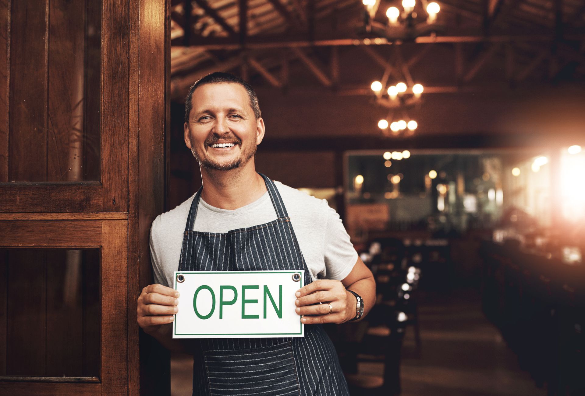 A man in an apron is holding an open sign in front of a restaurant door.