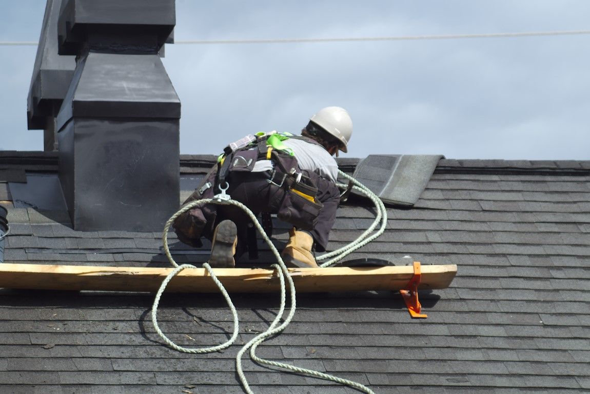 Roofer wearing safety harness working on a dark shingled roof near a chimney.