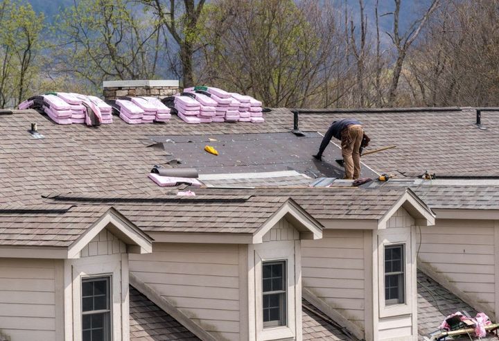 Roofer working on a brown shingled roof, next to stacks of pink insulation, against a background of trees.