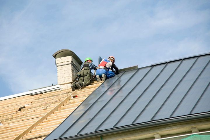 Two workers on a metal roof, one near a chimney, both wearing safety gear. Blue sky.