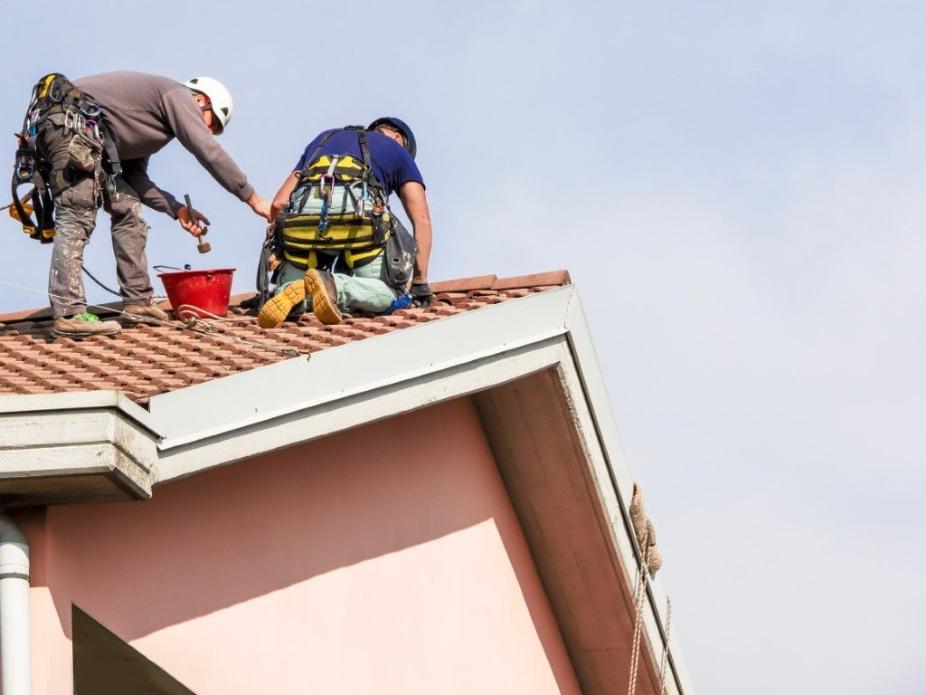 Two roofers, wearing safety harnesses, work on a red-tiled roof under a blue sky.