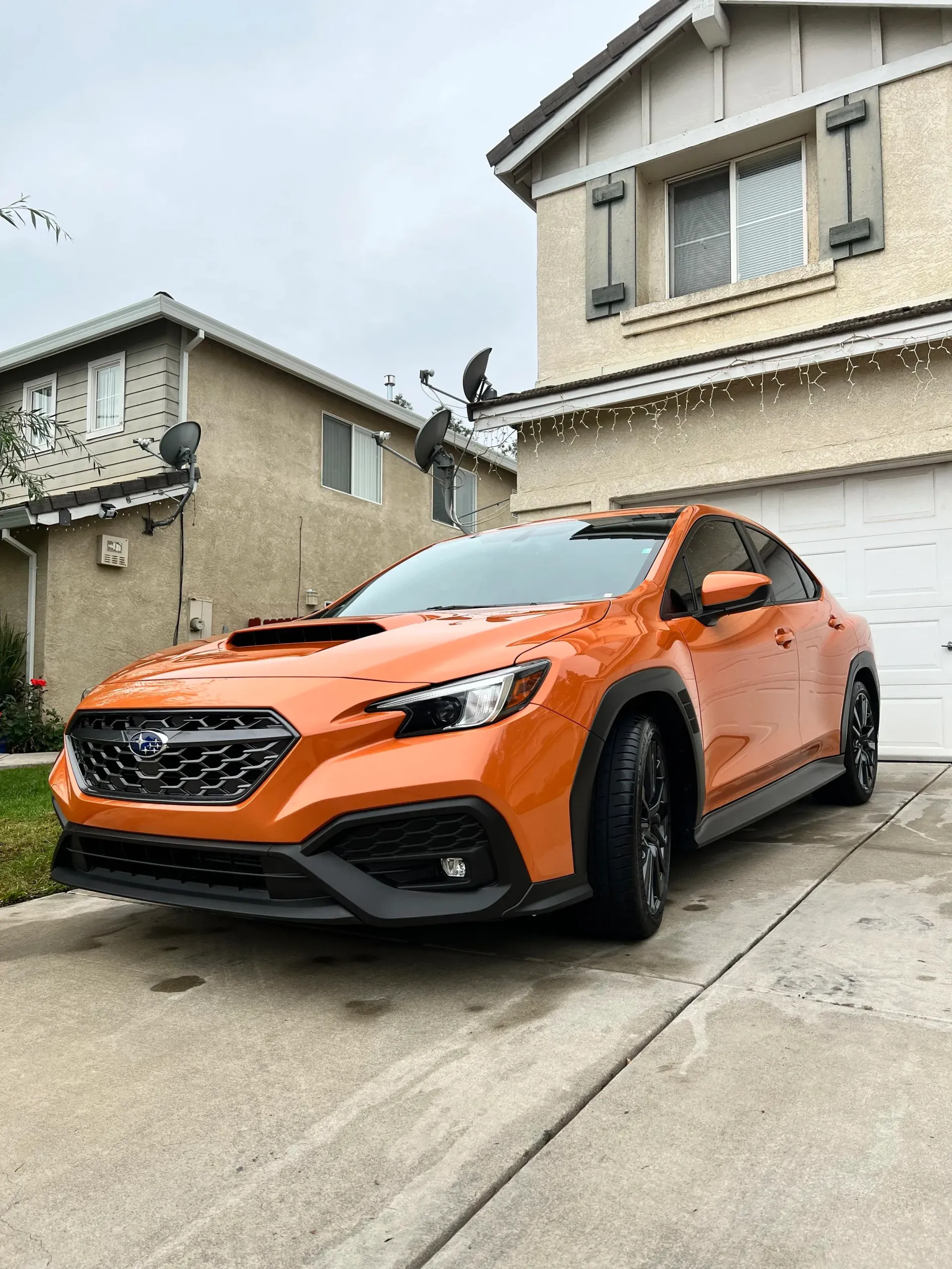 Orange Subaru WRX parked in front of a house on a driveway, cloudy sky.