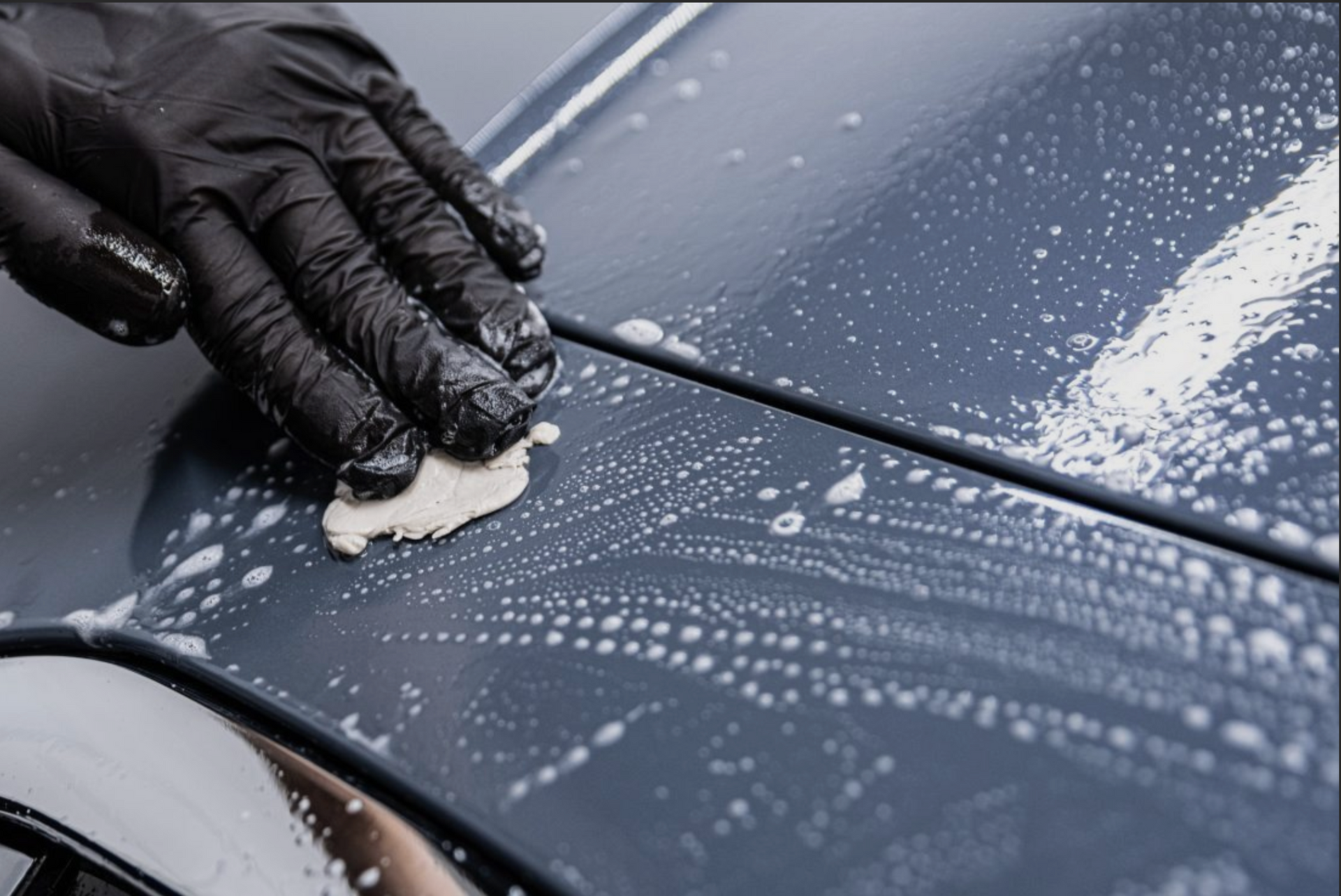 Gloved hand applying detailing clay to a car's surface, creating a soapy, textured effect on the blue paint.